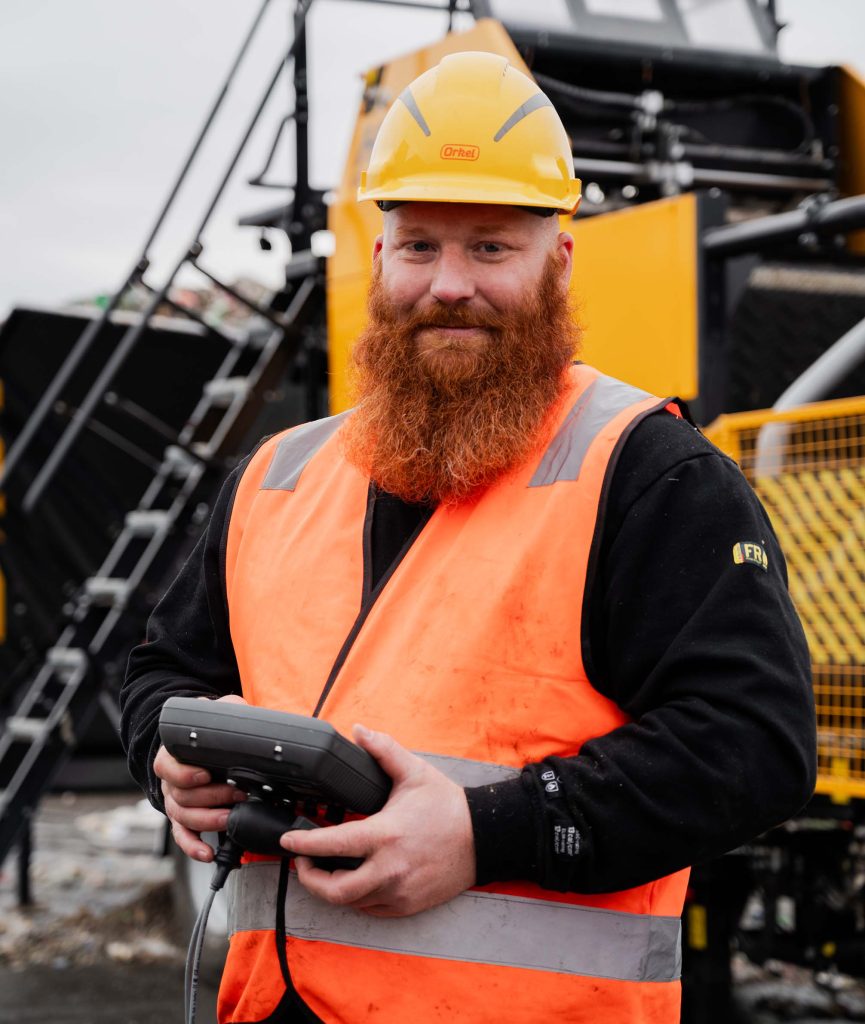 A man in a safety vest and helmet stands confidently in a waste facility, with a large industrial machine in the background. The setting includes piles of recyclable materials and heavy-duty equipment, emphasizing an industrial environment focused on waste management and processing.
