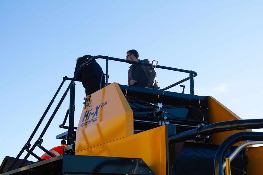 Two men standing on the platform of a industrial baler, looking away from the camera.