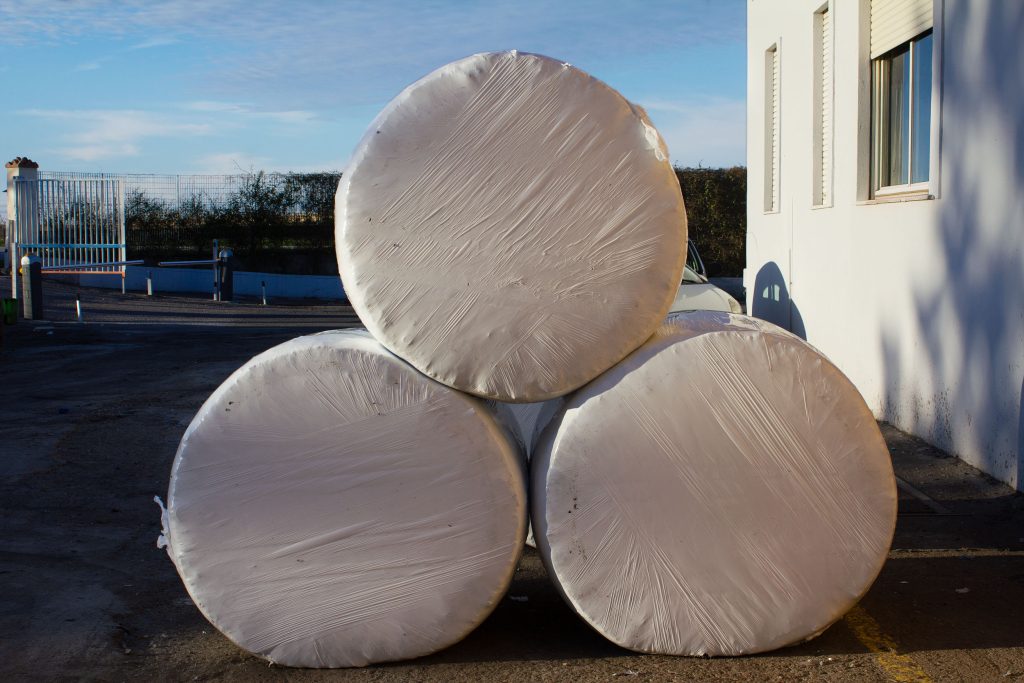 Three round bales wrapped in white plastic, stacked neatly beside a white building.