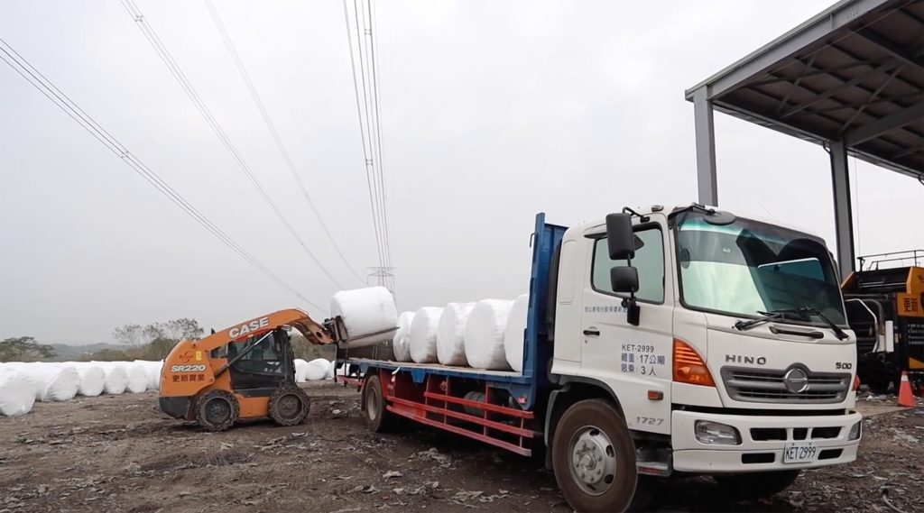 A forklift loading round bales onto a truck for transport, outside a large shed