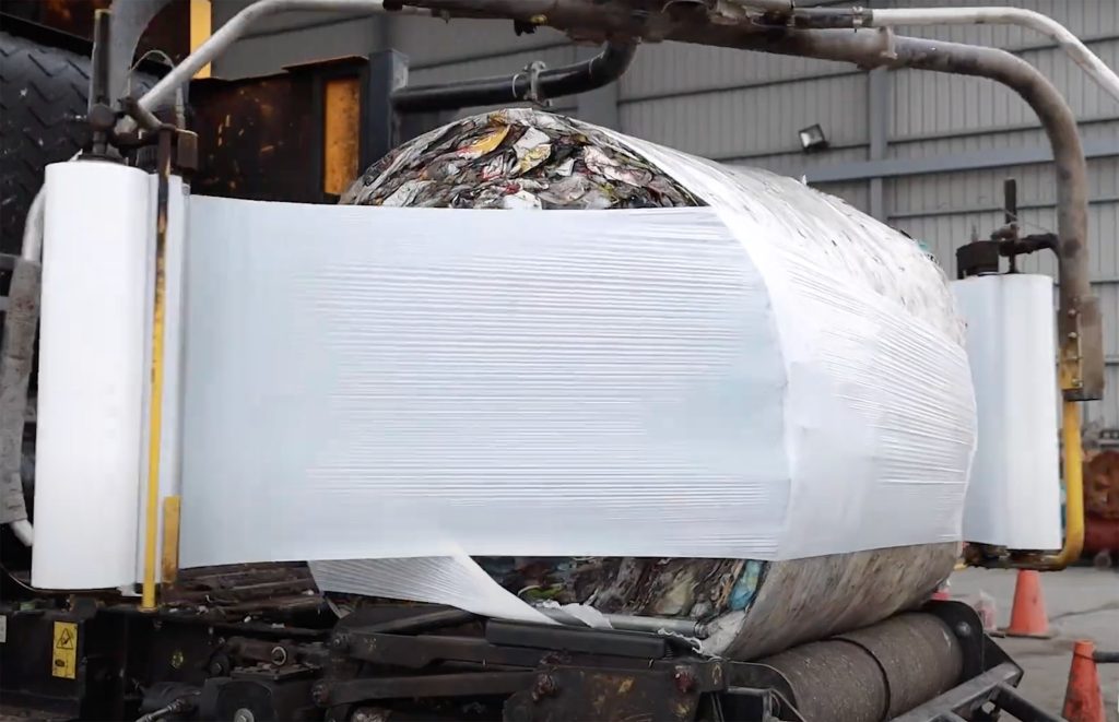 Closeup of a round bale of shredded waste being wrapped in plastic with an industrial baler