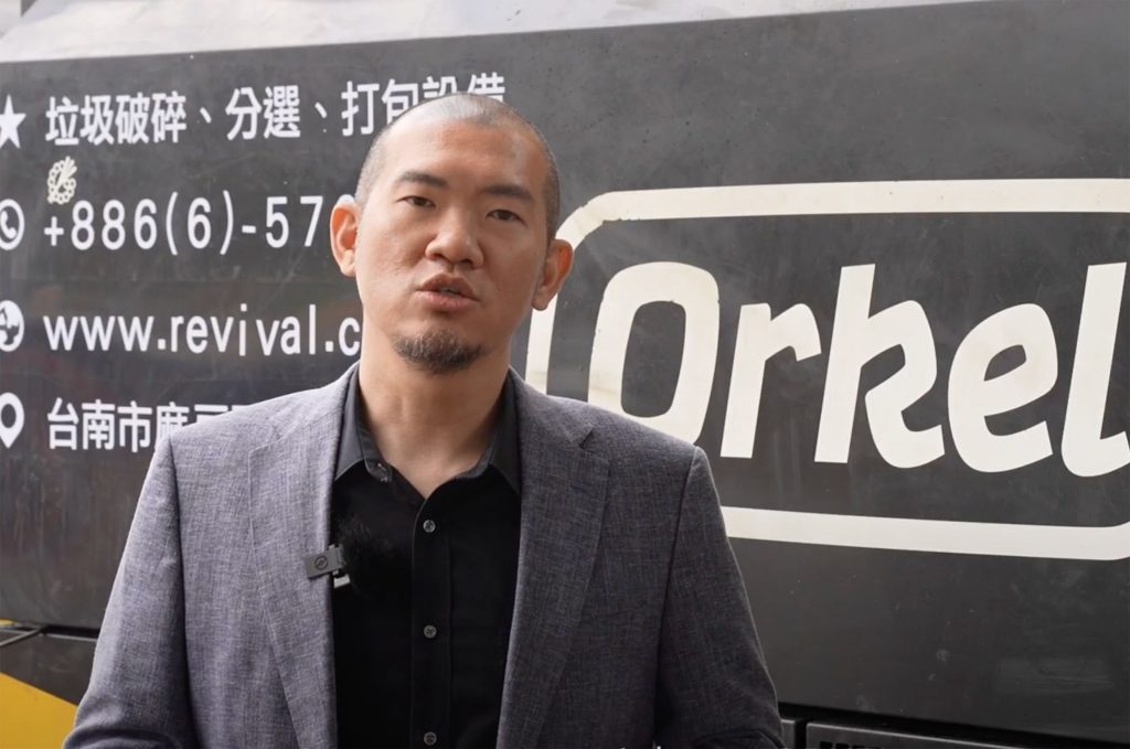 A man being interviewed, wearing a suit and a black polo shirt. An industrial machine in the background
